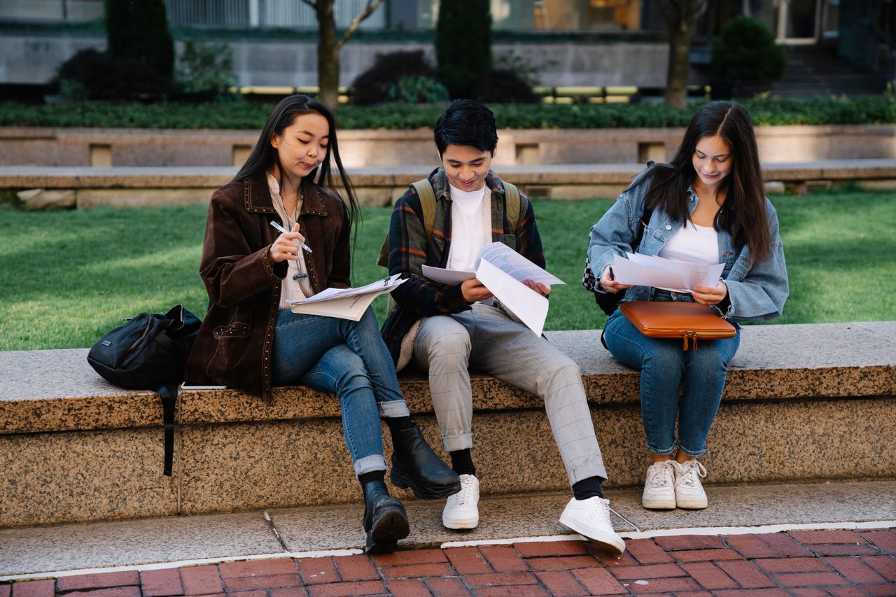 Three students studying together on a bench in a university campus setting outdoors.